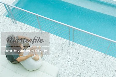 Husband and wife sitting by pool, his arm around her shoulder, her head on his