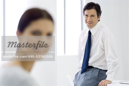 Professional man leaning against desk, smiling at camera, female colleague in foreground