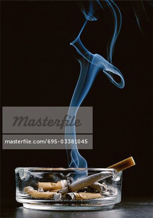 Ashtray with cigarette butts and lit cigarette smoking against black background