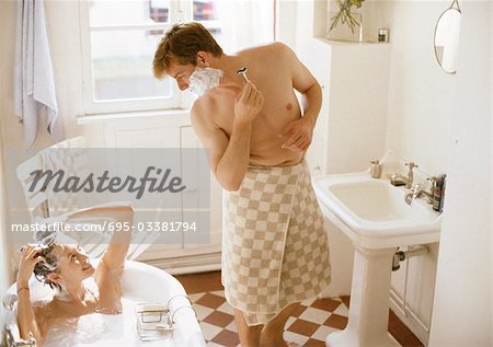 Woman taking bath, man with towel wrapped around waist shaving, in bathroom