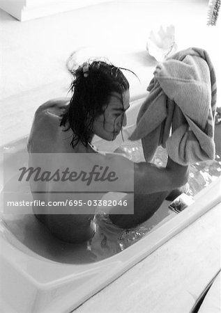 Long haired man sitting in bathtub, holding towel, rear view, high angle view, b&w