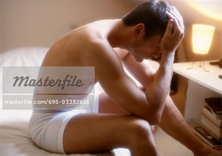 In bedroom, man sitting on bed in under garments, holding head