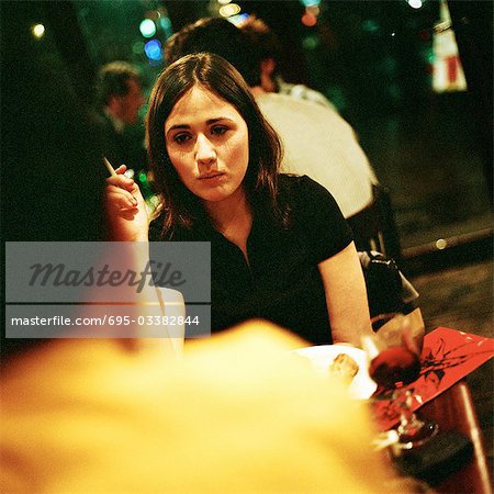 Young woman sitting at table and smoking, second person blurred in foreground