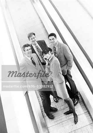 Group of business people on moving walkway smiling at camera, business woman in front on cell phone, b&w.