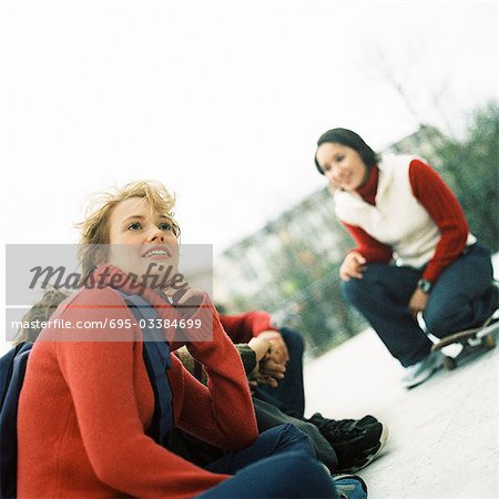 Young people outdoors, one with knee on skateboard