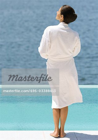 Woman in bathrobe standing near pool overlooking sea, rear view