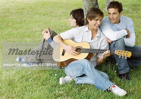 Junge männliche Freunde sitzen in der Nähe von Baum, Gitarre spielen