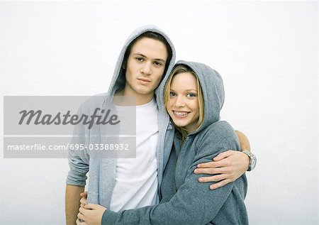 Young couple standing with arms around each other, wearing hooded sweatshirts, looking at camera