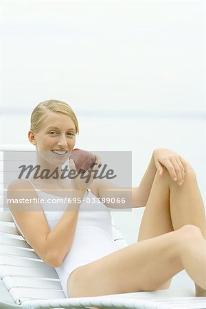 Teenage girl in swimsuit sitting on lounge chair, holding apple