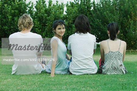 Groupe de jeunes amis assis sur l'herbe, vue arrière, une jeune femme regarder par-dessus l'épaule à la caméra