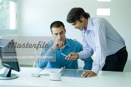 Two young businessmen in office, using cell phone, looking down