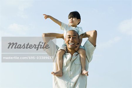 Father carrying son on shoulders and smiling at camera, child pointing and looking away