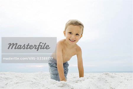Petit garçon accroupi sur la plage, souriant à la caméra, portrait