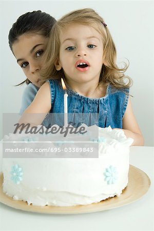 Girl blowing out candle on birthday cake, sister peeking at camera from behind