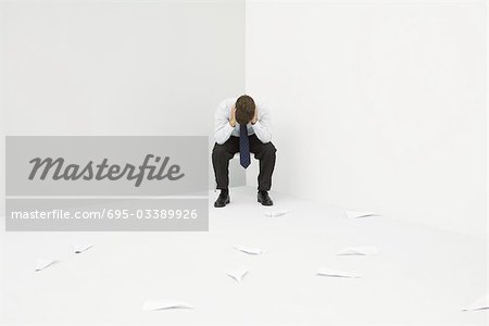 Professional man sitting in corner with head down, paper airplanes littering the floor