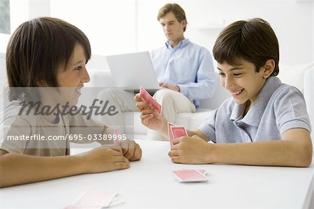 Brothers playing card game at table, father sitting on couch with laptop in background