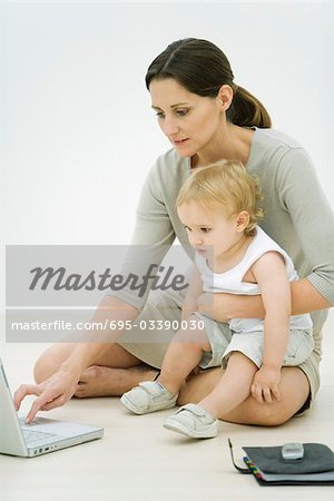 Professional woman sitting on the ground, using laptop, holding toddler on lap