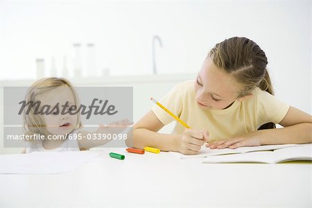 Girl doing homework at table, younger sister reaching for crayons