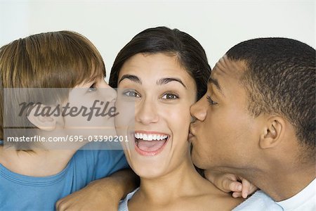 Woman smiling at camera as husband and son kiss her cheeks