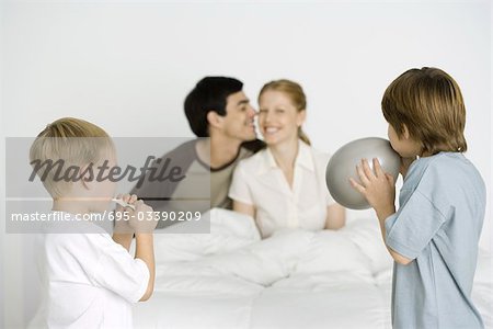 Two boys inflating balloons, parents smiling on bed in background