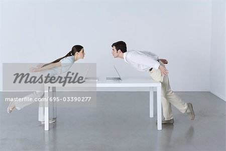 Man and woman at opposite ends of table, leaning towards each other, laptop computers between them