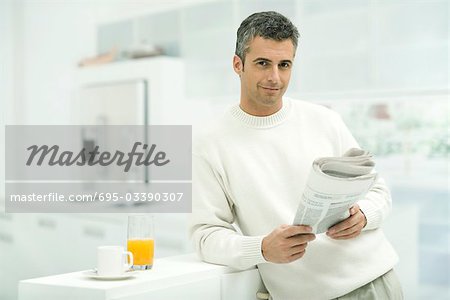 Man leaning against kitchen counter, holding newspaper, smiling at camera