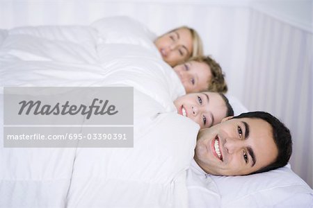 Family lying together in bed under comforter, smiling at camera