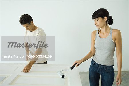 Woman painting door with paint roller, man protecting woodwork with masking tape