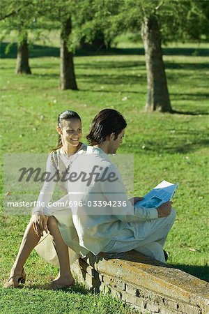 Couple sitting outdoors on low wall, man reading newspaper