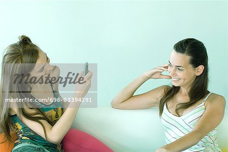 Two young friends sitting on couch, one taking photo of the other with cell phone