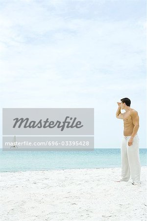 Man standing at the beach, looking at sailboat in the distance, side view