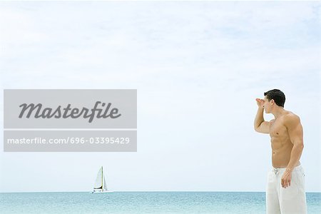 Man standing at the beach, looking at view, sailboat in the distance