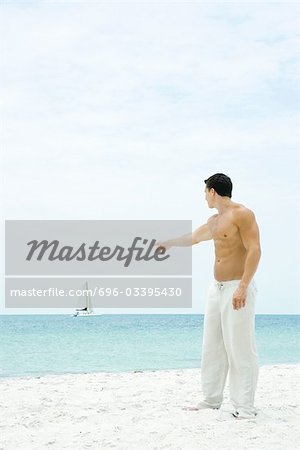 Homme debout sur la plage, en pointant sur le voilier au loin, vue latérale