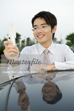Young businessman leaning against car, holding up cell phone, smiling