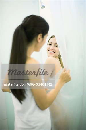 Young woman taking shower while friend holds open shower curtain