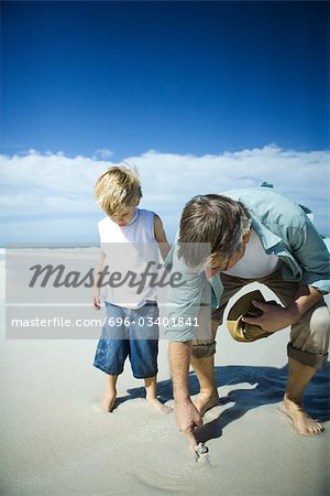 Senior man and grandson on beach, man sticking finger into sand