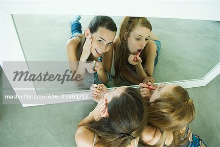 Two friends lying on ground, applying lipstick, looking in mirror, one looking at camera, view from directly above