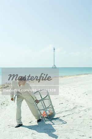 Man in suit pulling garbage can across sunny beach, looking back over shoulder, full length