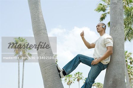 Man wearing headphones leaning against tree with thumb raised, looking away