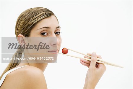 Young woman eating grape with chopsticks, looking over shoulder at camera