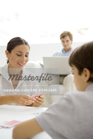 Woman playing a card game with her son, husband sitting in background with laptop