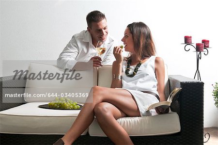 Young man leaning on couch with white wine and young woman reading book and eating grapes