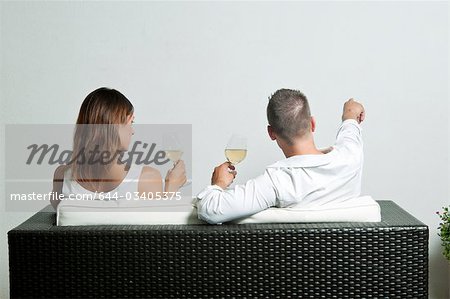 View of couple from the back sitting on couch drinking white wine, young man pointing