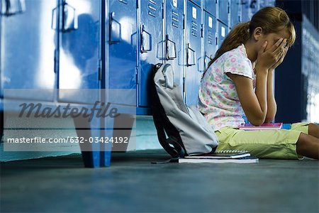 Female junior high student sitting on hall floor near lockers, upset and covering face with hands