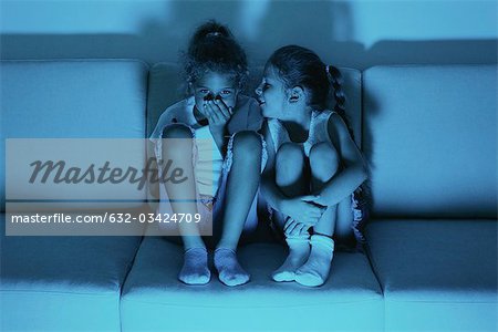 Two girls watching TV together, one whispering in the other's ear