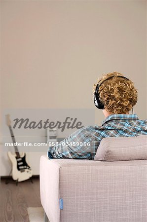Man with headphones sitting on a couch, e-guitar in the background, rear view