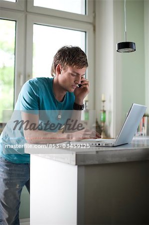 Jeune homme debout au comptoir et à l'aide de téléphone portable et ordinateur portable, faible angle vue