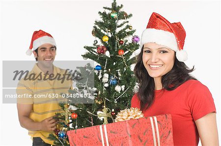 Woman holding a Christmas present with a man decorating a Christmas tree in the background