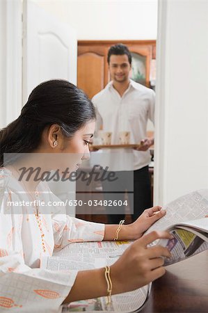 Woman reading a newspaper with her husband bringing coffee