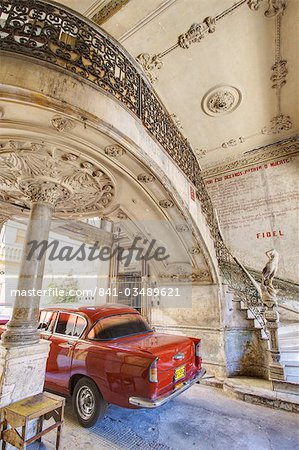 Classic red American car parked beneath ornate marble staircase inside dilapidated apartment building, Havana, Cuba, West Indies, Central America